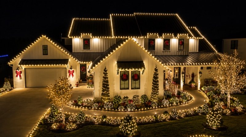 White farmhouse fully outlined in warm white lights with lit wreaths, bows, and a landscaped light display